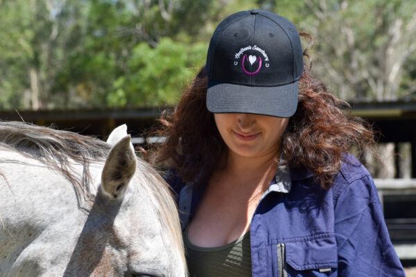 A model posing with a black Hoofbeats Cap next to a horse